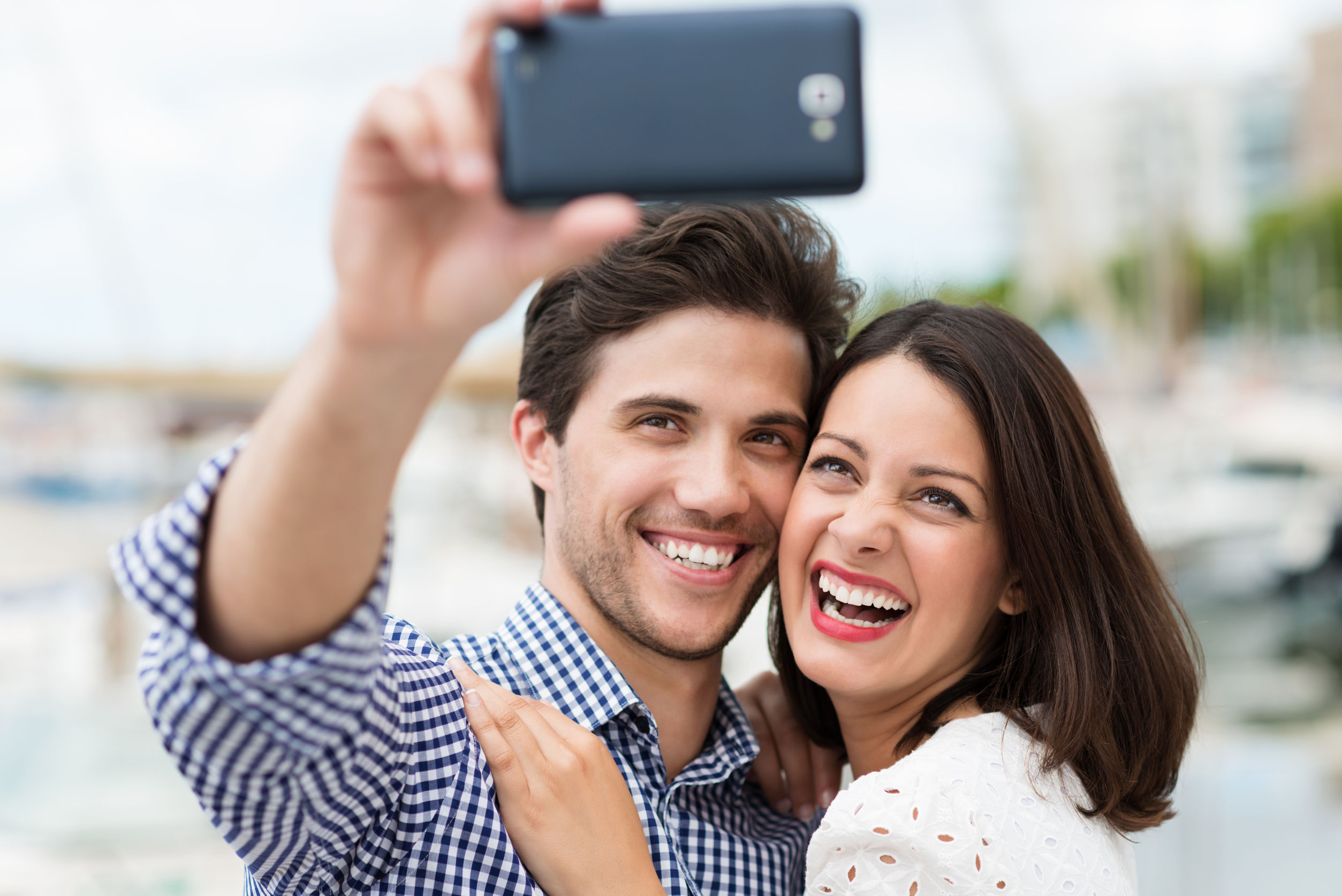 young couple taking a self portrait laughing as they pose close together for the camera on their smartphone outdoors in summer sunshine