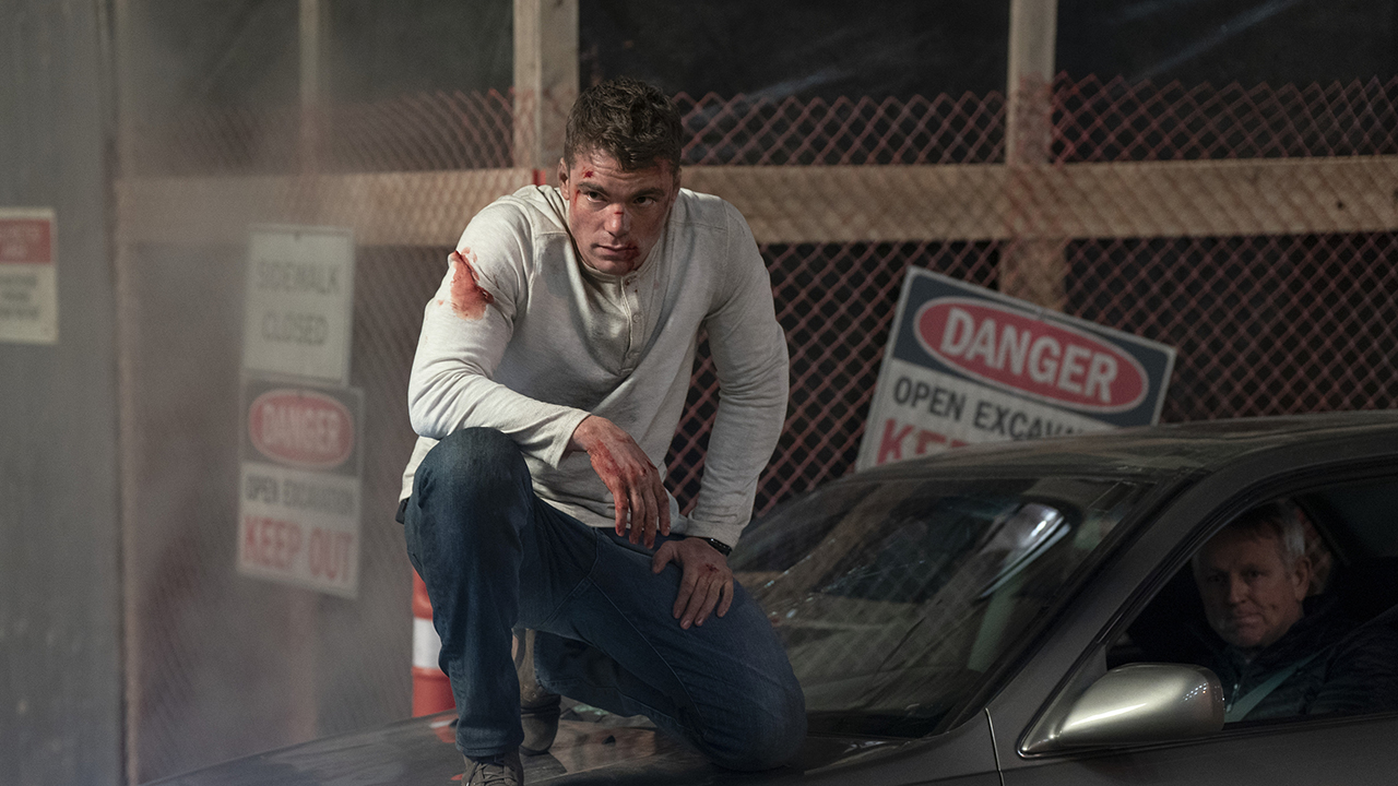 Peter kneeling on top of a car, bloody and battered in a scene from The Night Agent.