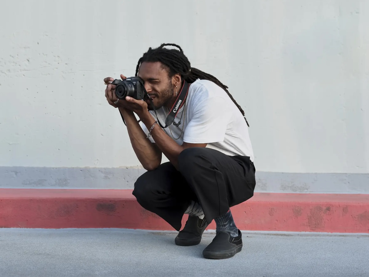 A man kneels down on the sidewalk to take a picture with the Canon EOS R7 mirrorless camera.