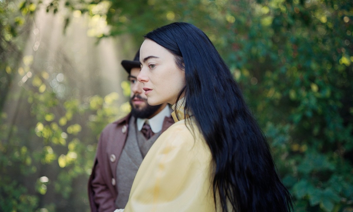 Emma Stone stands in front of Ramy Youssef in a still from Poor Things