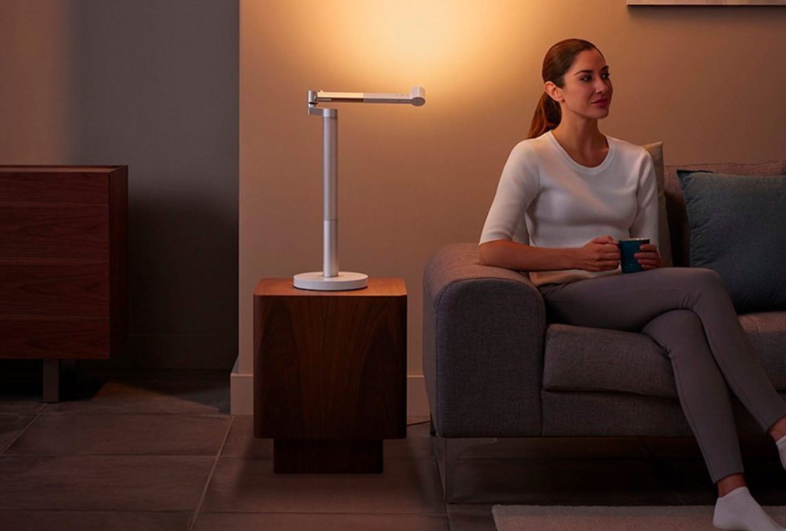 A woman sits on a couch nexts to the Dyson Solarcyle Morph smart desk light.