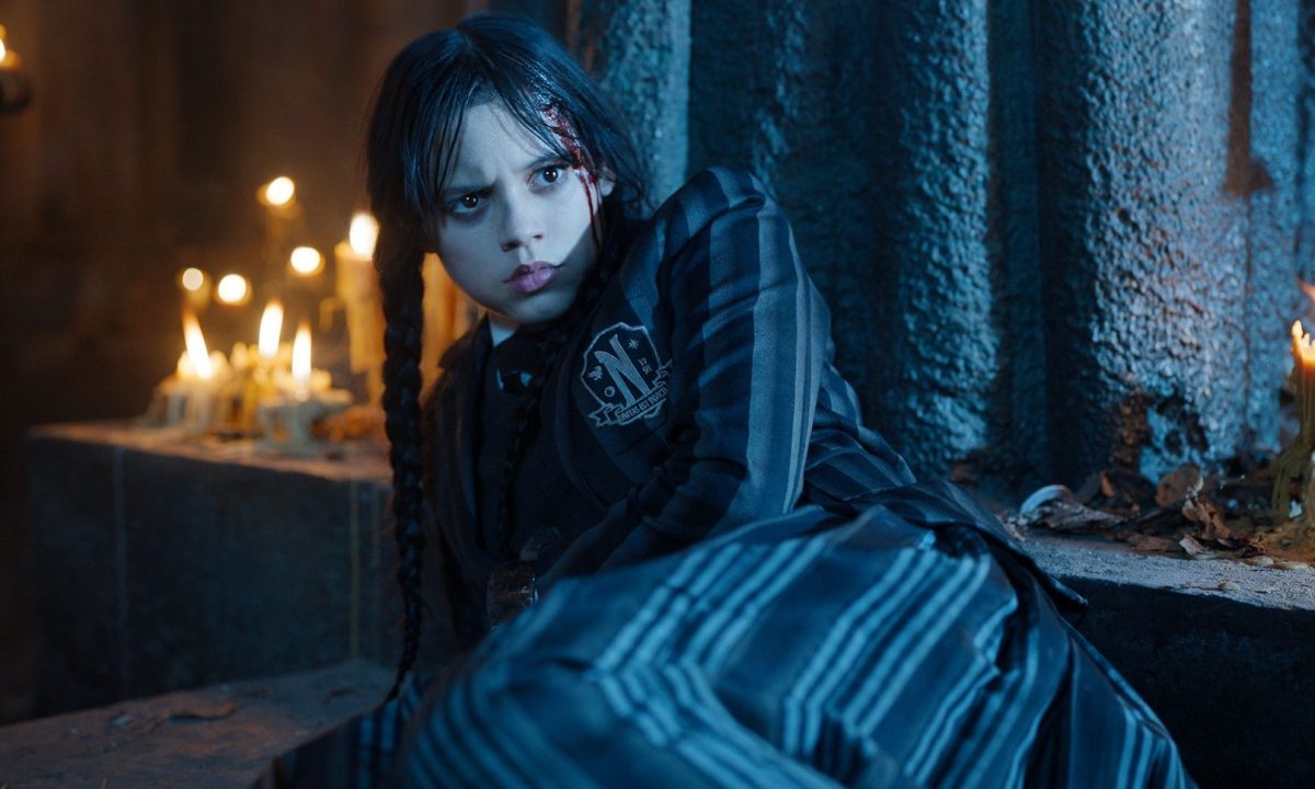 Jenna Ortega lays on the ground and stares over her shoulder.